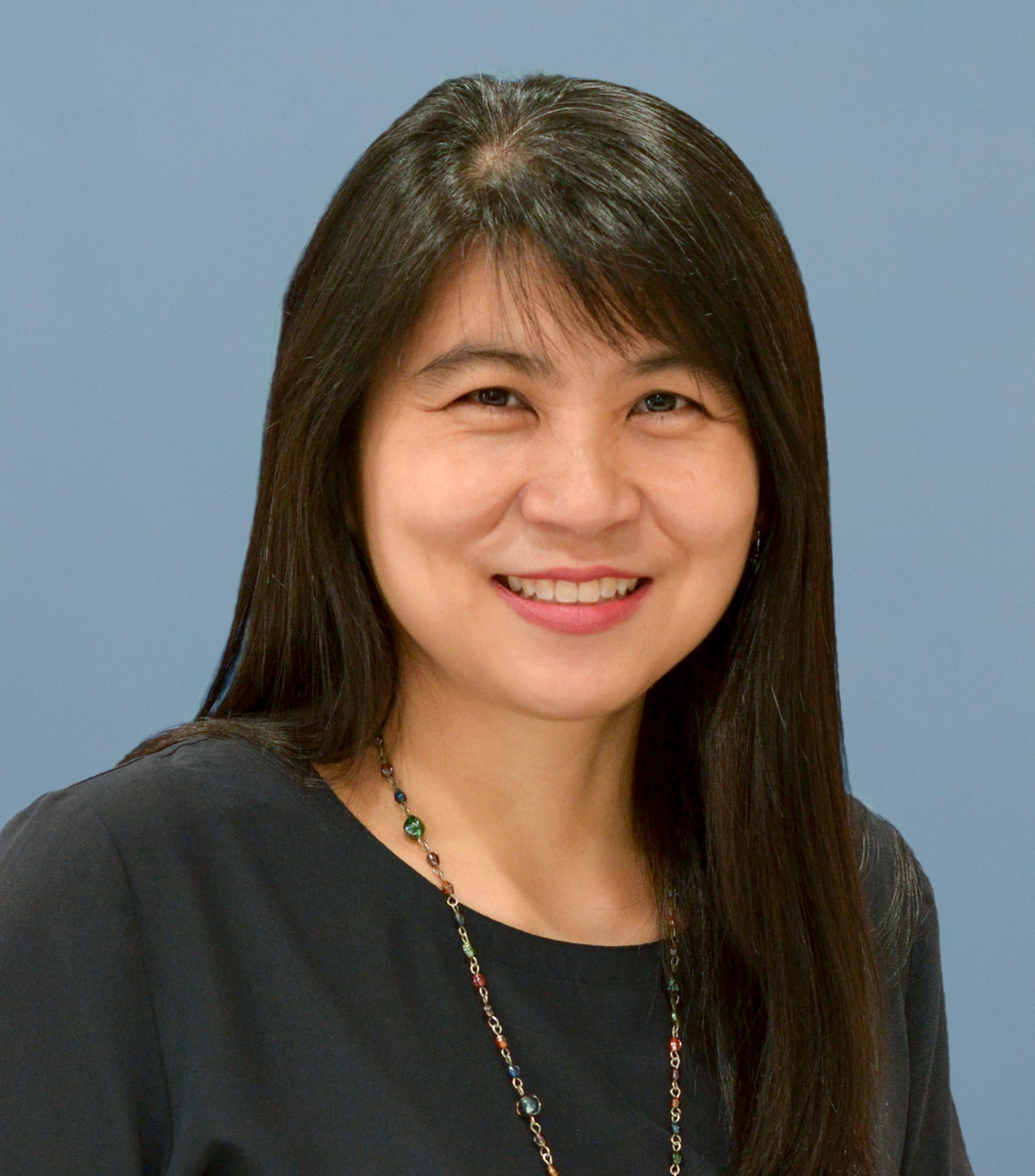 Woman with long dark hair smiling, wearing a black top and colorful beaded necklace against a blue-gray background.